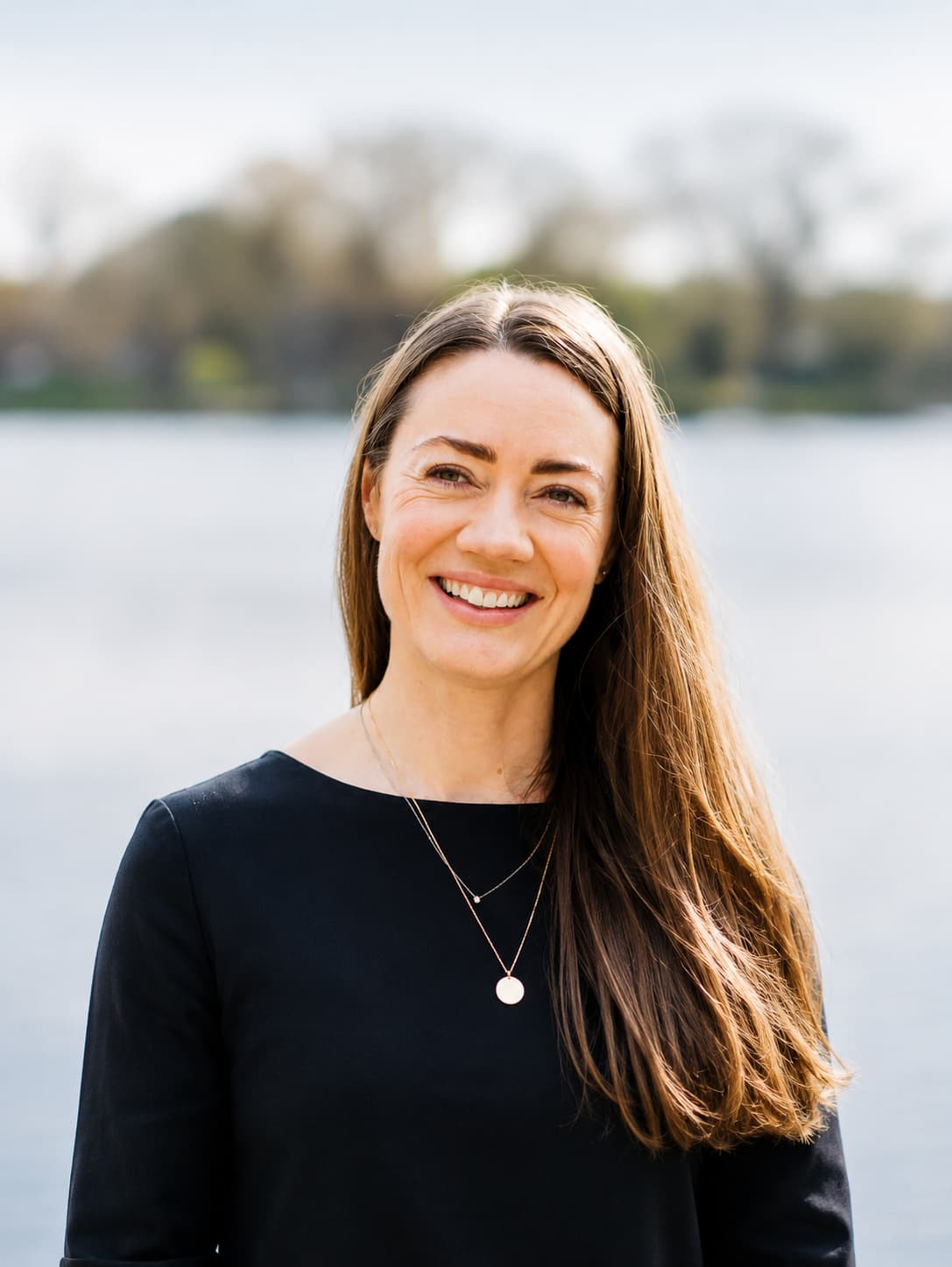 Smiling woman with long brown hair in a black top, standing by a lake with blurred trees in the background.