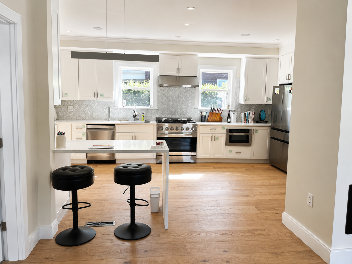 Bright white kitchen with an island, two black bar stools, and stainless steel appliances. It shows a modern, airy space with light wood floors.