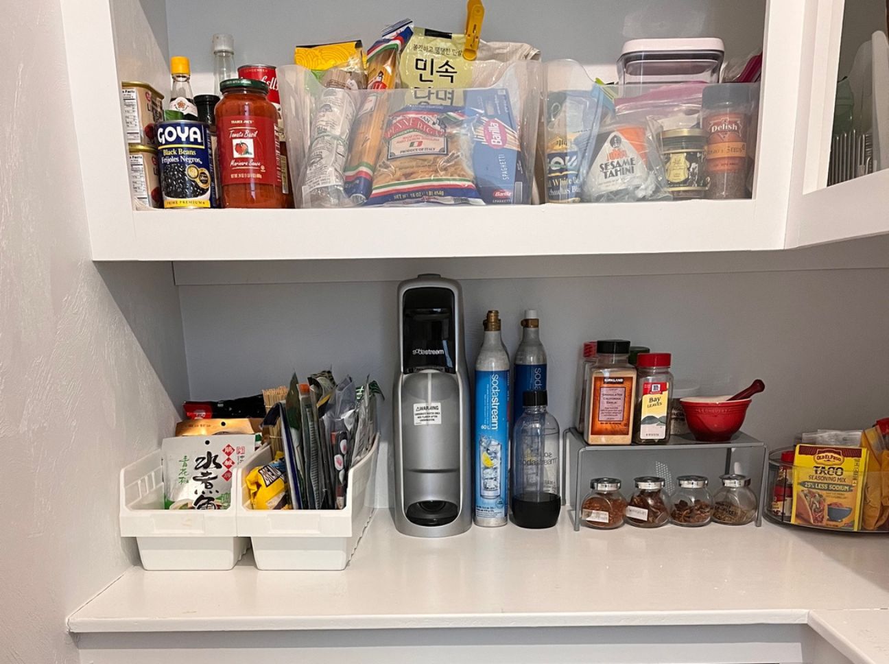Organized kitchen cabinet with a coffee station: Nespresso-style machine, pods, spices, and labeled containers on the counter; snack bins on the upper shelf.
