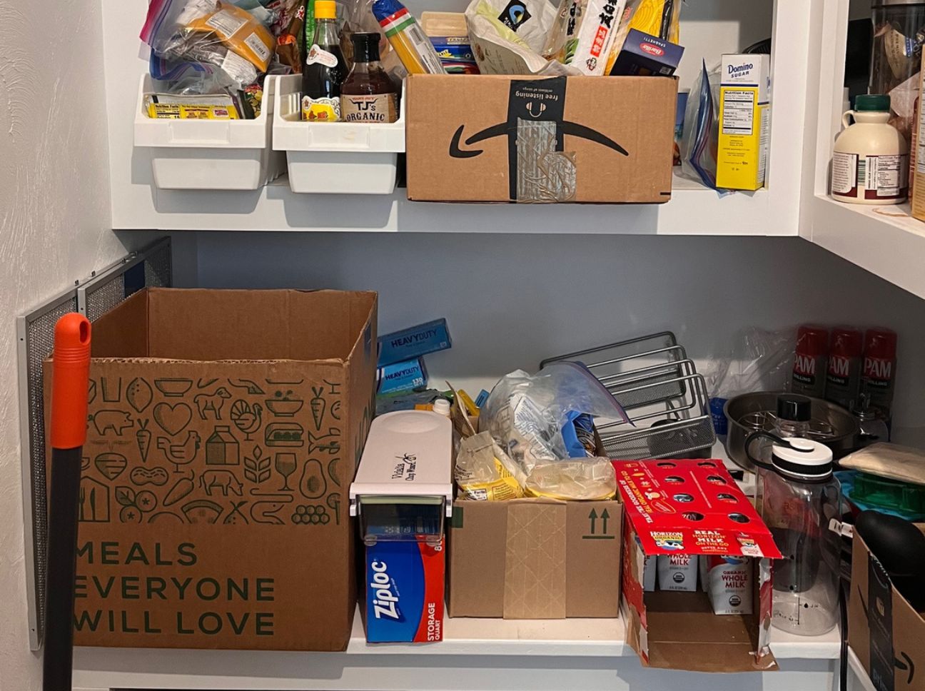 Cluttered pantry shelf with cardboard boxes and grocery items, including a large 'Meals Everyone Will Love' box.