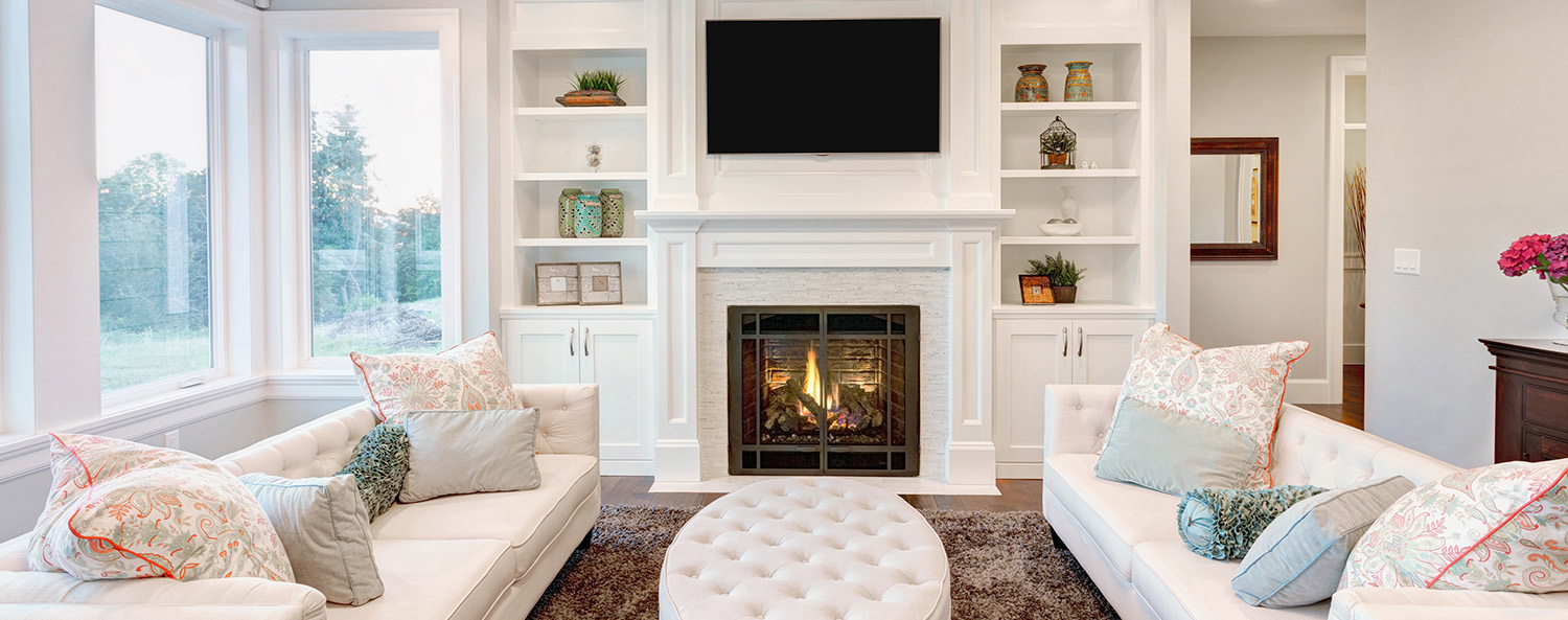 A cozy living room with two white sofas facing each other, adorned with colorful pillows. A round, tufted ottoman sits between them. A fireplace with a TV above it is centered on the back wall, flanked by built-in shelves with decorative items. Large windows provide natural light.