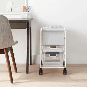 A white rolling cart with mesh drawers is placed next to a gray office chair and dark wood desk. The cart has three storage tiers with various office supplies, including files on the top tray. The desk has a mug filled with pencils and other writing tools.