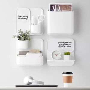 A wall organizer with four white compartments, holding scissors, pens, a potted plant, cards, a clock, and letters. Two compartments have whiteboards with reminders. A desk below has headphones, a laptop, and a coffee cup.