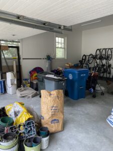 A messy garage with recycling bins, bags, and various items like bicycles on the wall, garden tools, and paint cans. The space has a concrete floor and a window letting in natural light. A paper yard waste bag is in the center.