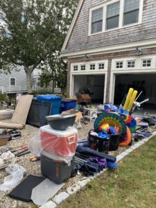 A garage organizing scene with various items spread outside a garage. Items include colorful pool floats, plastic bins, a vacuum, and a cooler, all set on gravel. The garage itself is open, with more items visible inside. Trees and houses are in the background.