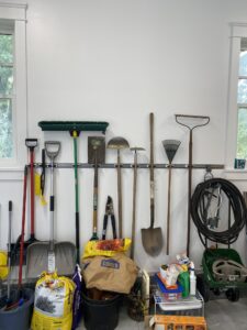 A garage wall with various garden tools hanging, including shovels, a broom, and rakes. Below, bags of garden soil and seeds are stacked, alongside a hose and a small cart with miscellaneous items. Two windows frame the scene.