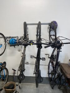 Three bicycles are mounted vertically on a wall rack in a garage. Bike helmets rest on one of the handlebars. A wooden table and parts of additional bikes are visible to the sides. A scooter and a basket are on the floor.