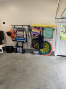 A neatly organized garage wall with beach chairs, a colorful float, and a pool noodle hanging. Below are a beach cart and a plastic storage container. The garage floor is clean, and a door with windows offers a view of the outside.