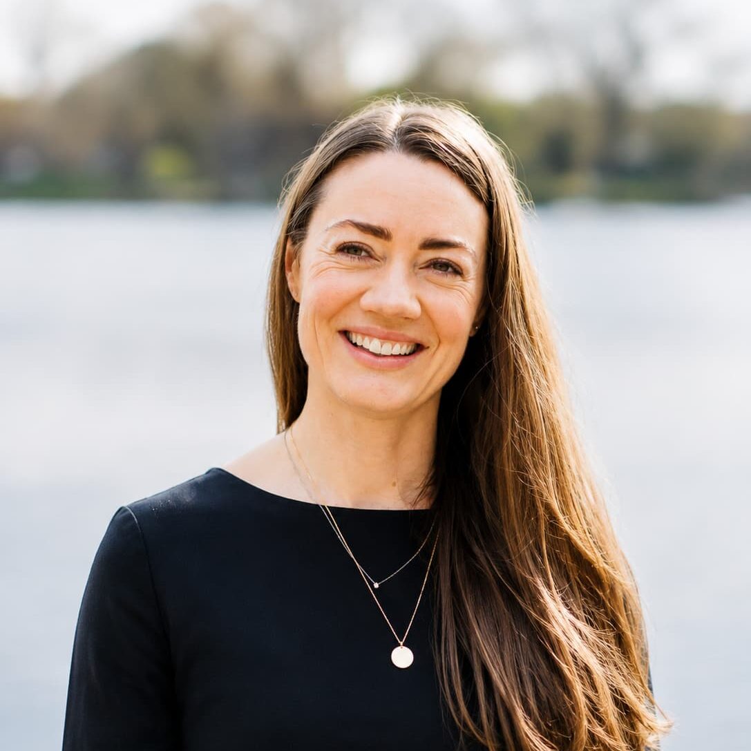Smiling woman with long brown hair in a black top, standing by a lake with blurred trees in the background.