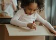Child drawing on a large cardboard moving box with a red marker, concentrating on a craft project.
