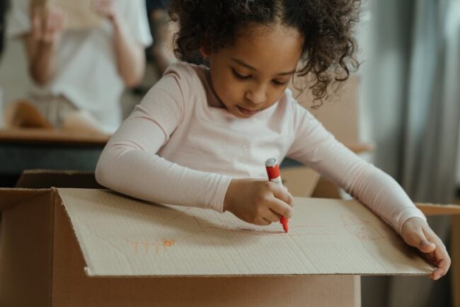 Child drawing on a large cardboard moving box with a red marker, concentrating on a craft project.
