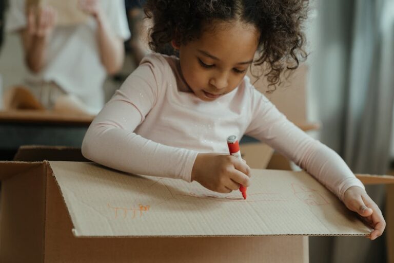 Child drawing on a large cardboard moving box with a red marker, concentrating on a craft project.