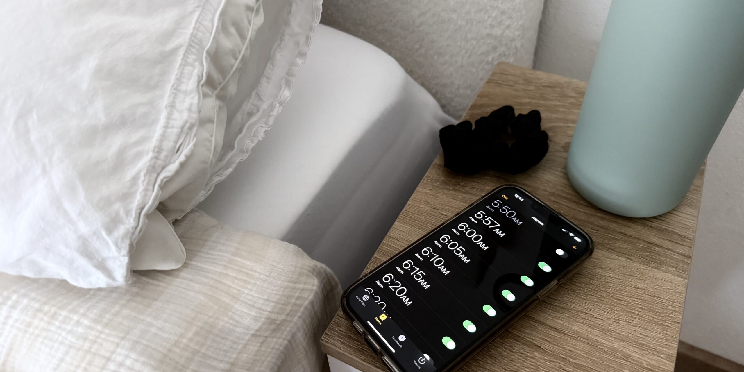 paige-blackmon-dupe Bedside scene with a wooden nightstand, a teal lamp, and a smartphone showing alarm times on screen as alarms glow.