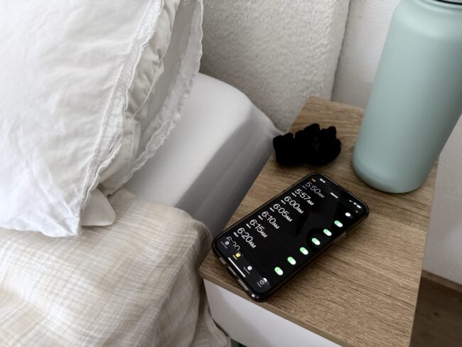 paige-blackmon-dupe Bedside scene with a wooden nightstand, a teal lamp, and a smartphone showing alarm times on screen as alarms glow.