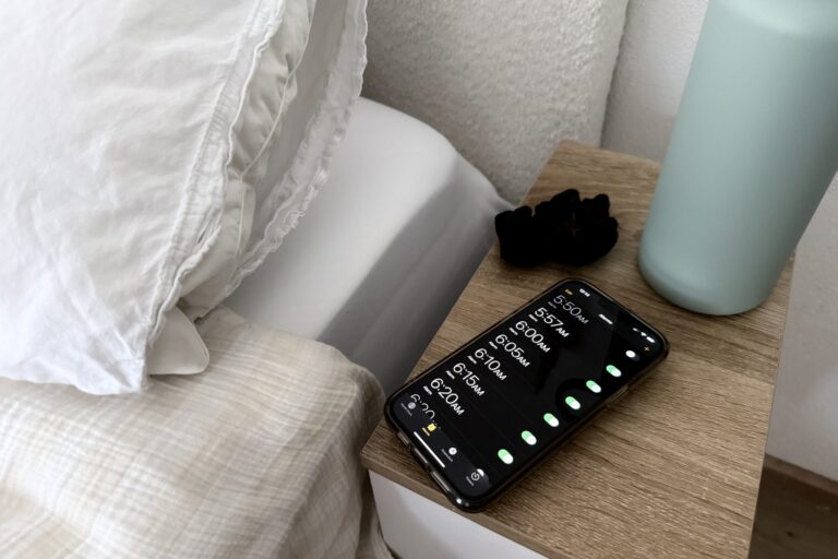 paige-blackmon-dupe Bedside scene with a wooden nightstand, a teal lamp, and a smartphone showing alarm times on screen as alarms glow.