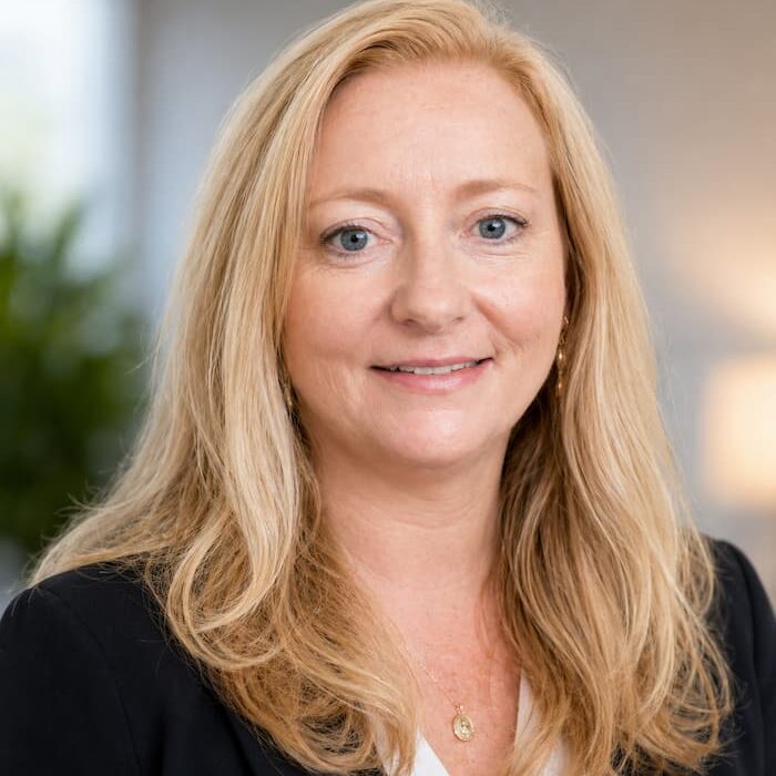 Professional headshot of a smiling blonde woman in a black blazer and white blouse, in a neutral office setting.